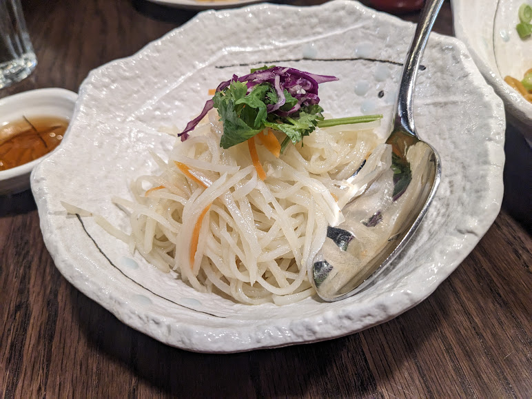 A bowl of thinly sliced white radish and carrot garnished with cilantro and purple cabbage, with a spoon resting in the bowl.