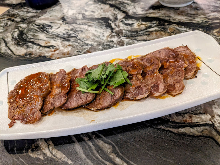 A plate of sliced, cooked beef garnished with cilantro served on a white rectangular dish, placed on a marble-patterned surface.