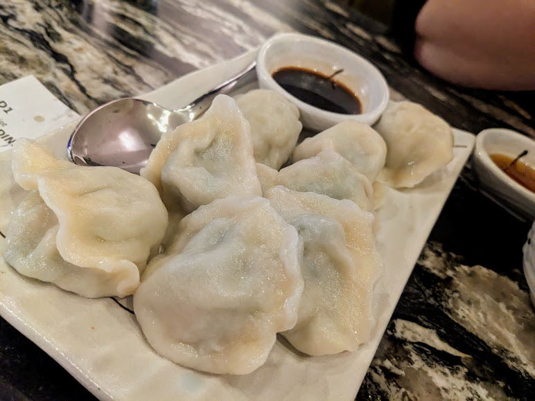 A plate of steamed dumplings served with soy sauce on a marble-patterned table. A spoon is placed on the plate alongside the dumplings.