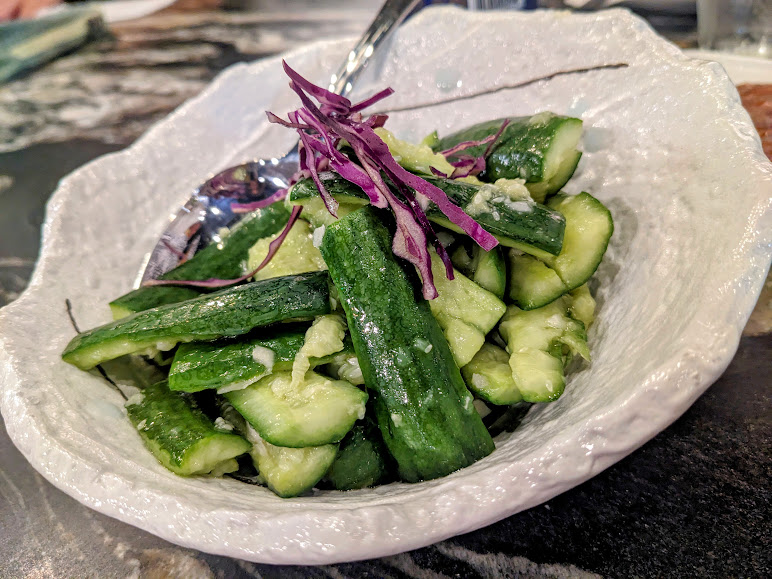 A white bowl filled with cucumber sticks, garnished with slices of purple cabbage, and a silver spoon resting in the bowl.
