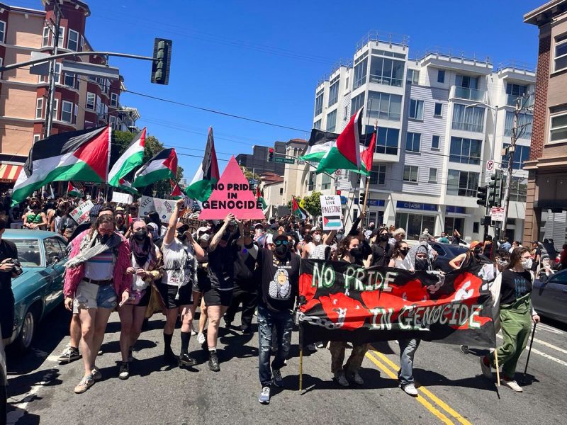 A group of people march on a city street holding Palestinian flags and signs with messages, including "No Pride in Genocide.