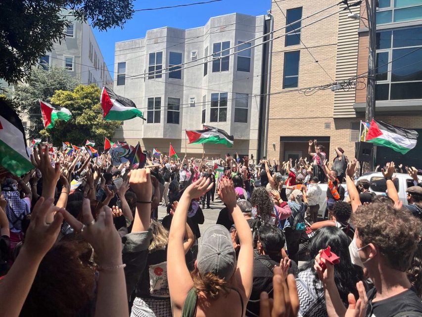 A large crowd of people with raised fists waves Palestinian flags in the street in front of apartment buildings during a sunny day protest.