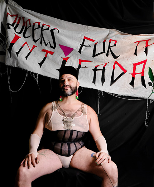 Mama Ganuush, a local Palestinian drag artist and activist, called for the boycott. They sit in front of a banner with the words "Queers for the Intifada" wearing a sheer top, black headpiece, and colorful earrings. Pride weekend.
