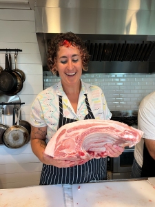 A person wearing a striped apron holds a large cut of raw meat in a kitchen with hanging pots and a stainless steel vent hood.