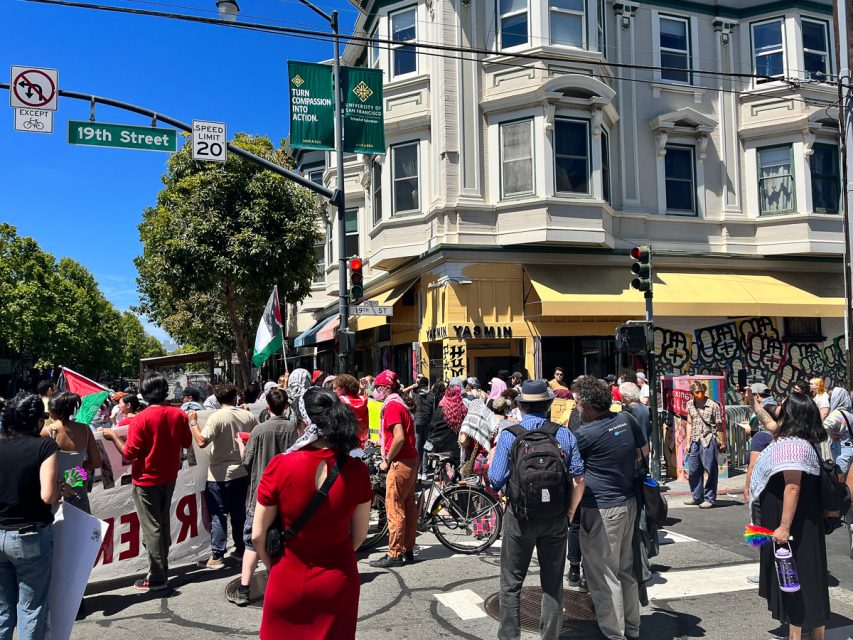 A group of people participate in a protest on a sunny day at an intersection near a building with a yellow awning. They hold signs and flags. The street signs read "19th Street" and "Mission Street. Vice President Kamala Harris was anticipated to arrive.