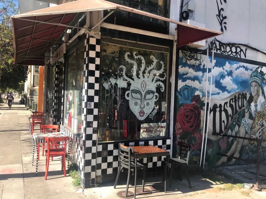 A corner cafe with checkered tiles, outdoor seating, and large murals on the walls. One mural depicts a face with white hair, while another features red roses and the word "Mission." Perfect for quick snaps over a coffee.