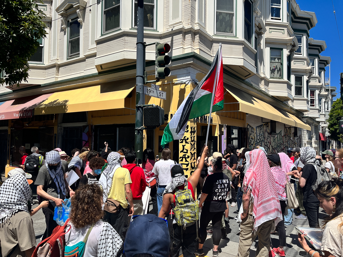 A group of people on an urban street corner march with flags and signs. Many wear checkered headscarves, and one carries a flag resembling the Palestinian flag. Storefronts and a traffic light are visible.