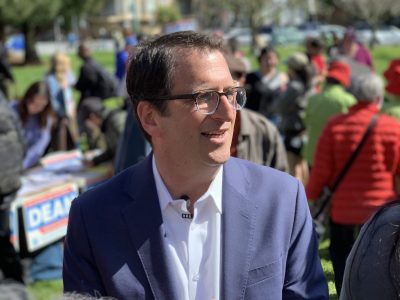 Dean Preston in glasses and a suit smiles outdoors at a District 5 event, with people and banners in the background.