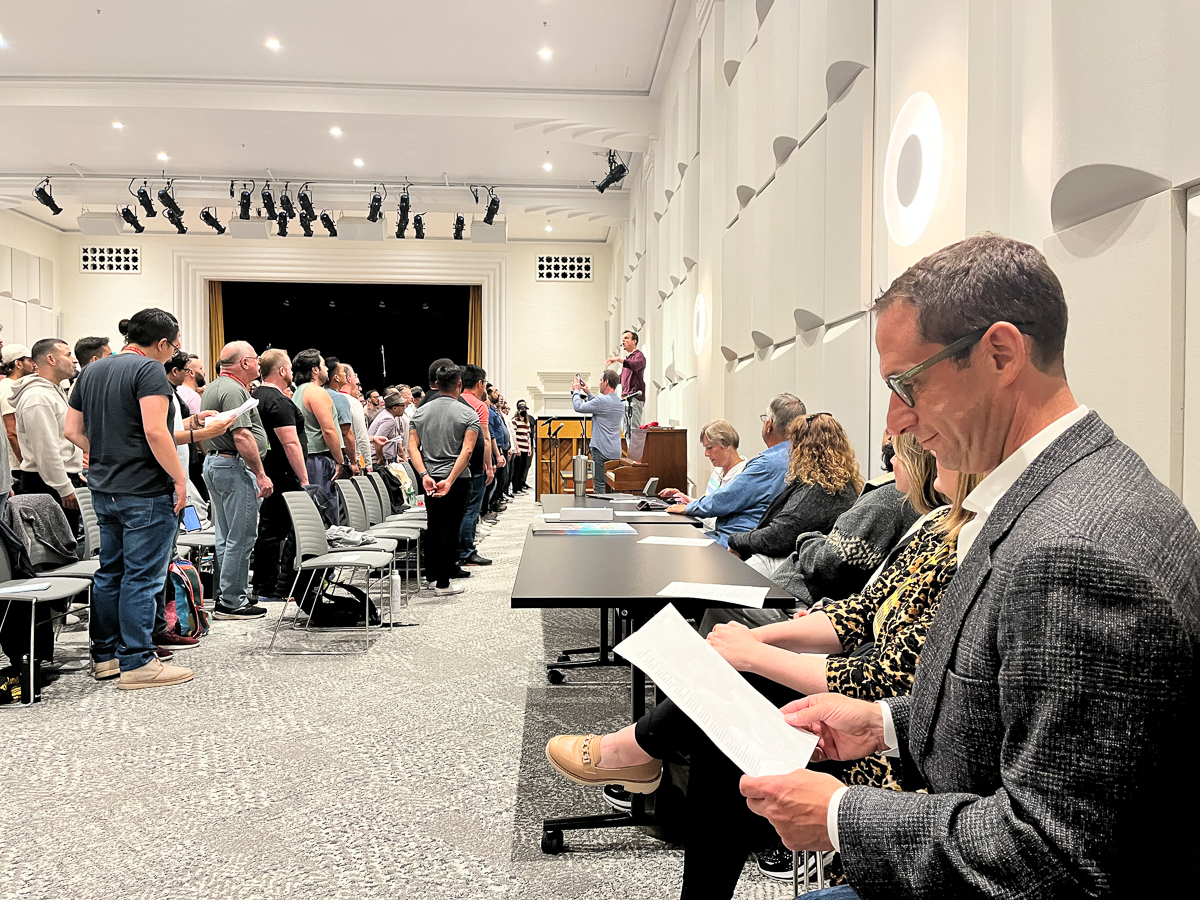 People in a conference room, some standing in line at the front, while others sit and listen. One person in the foreground is reading a paper.