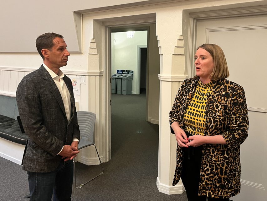 Two people having a conversation in an indoor setting. The man on the left is wearing a gray suit, the woman on the right is wearing a yellow and black patterned outfit.