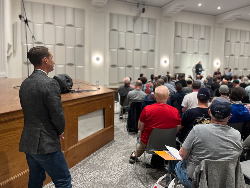 A man in a gray blazer stands near a stage, observing an audience seated in a room with white textured walls. Another man is speaking at a podium in front of the audience.