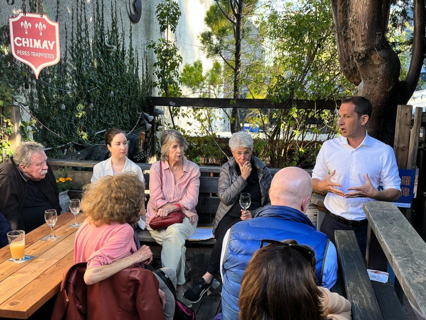 一群人坐在户外的木桌旁，一名男子站在一旁与他们交谈。桌子上放着饮料，背景是绿色植物。