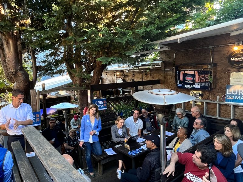 A person speaks to a small crowd seated outside on a wooden deck. They appear to be at a campaign event, with signs and buttons supporting "Lurie for Mayor" visible in the background.