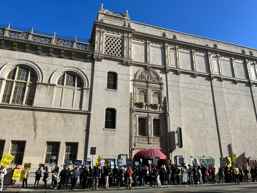 A group of people stands outside a large, ornate building, holding signs and banners.