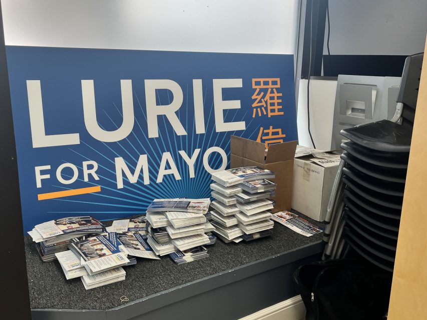 Stacks of campaign flyers and boxes are placed in front of a "Lurie for Mayor" sign next to a pile of black chairs.