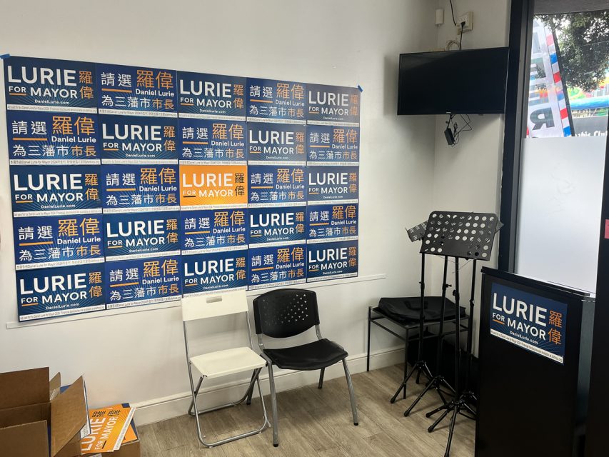 Campaign office with multiple "Lurie for Mayor" signs on the wall, two chairs, a podium, and a music stand inside. A glass door to the right shows a street view with some colorful elements outside.