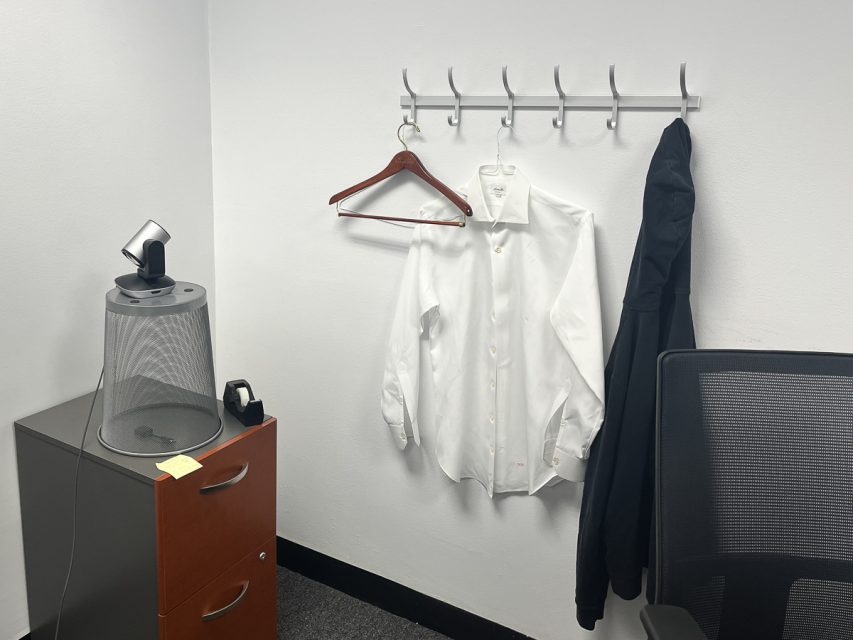 A white dress shirt and a black jacket hang on a wall-mounted rack next to an office chair and a small cabinet with a fan.