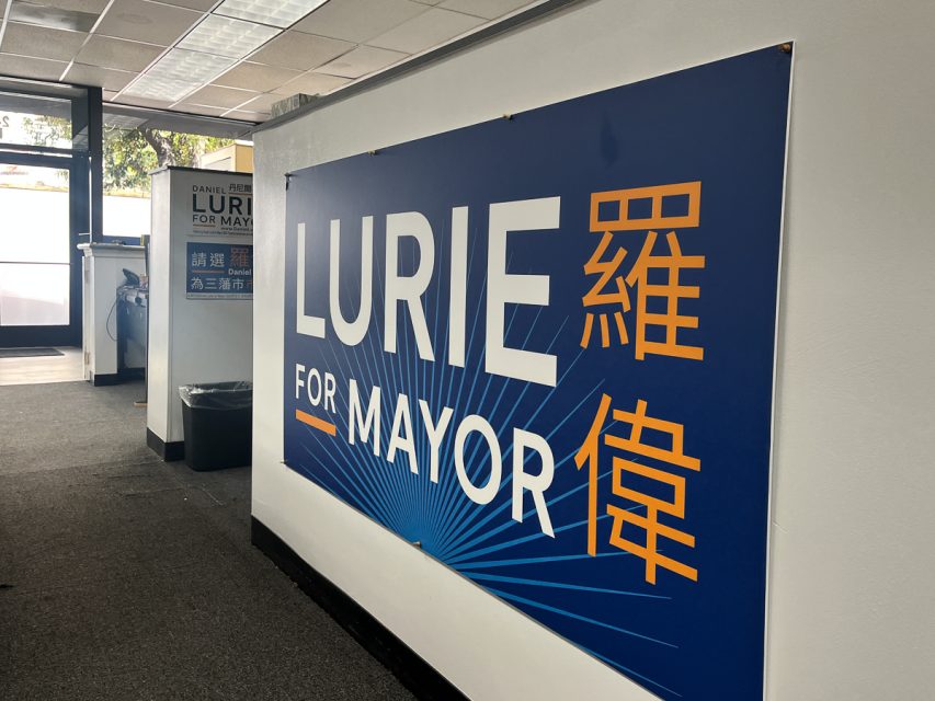 A campaign office interior featuring a large sign on the wall that reads "Lurie for Mayor" with additional text in another language.