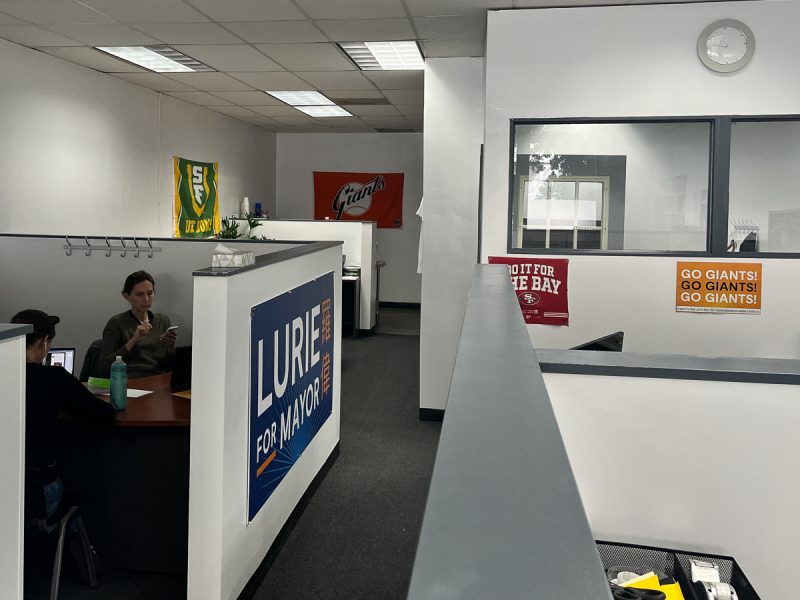 竞选办公室里有两个人在办公桌前办公，挂着 "罗伟竞选市长 "的大牌子，墙上挂着几个运动队的横幅。