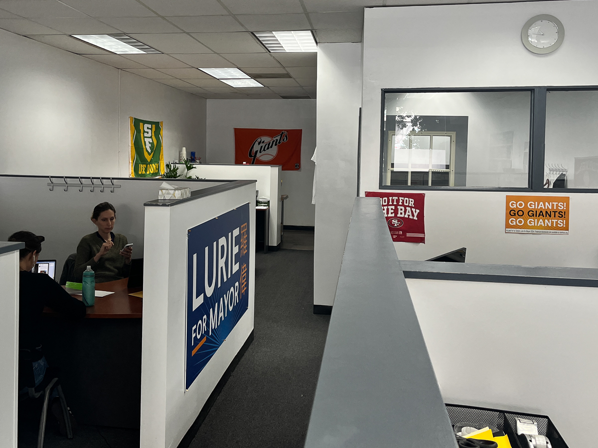 競選辦公室裡有兩個人在辦公桌前辦公，掛着 "羅偉競選市長 "的大牌子，牆上掛着幾個運動隊的橫幅。