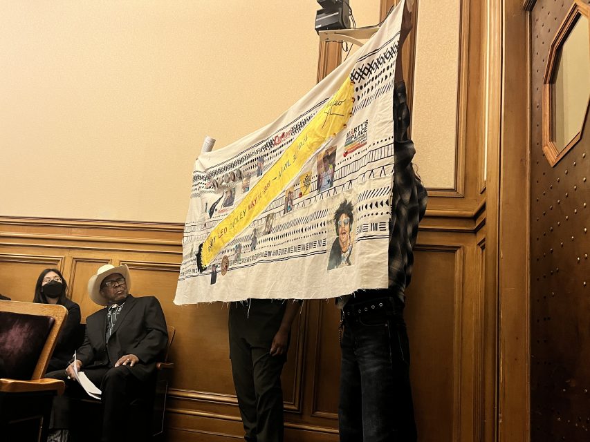 Two individuals hold up a large banner featuring images and text in a wood-paneled room. Seated behind them are two people, one wearing a hat and the other wearing a mask.