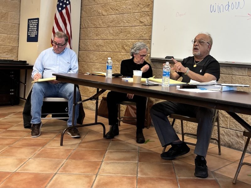 Three people sitting at a table with water bottles and papers during a meeting in an uptown office. An American flag and a whiteboard with the word "Window" are in the background.
