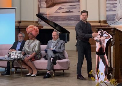 Four individuals are on a stage: one man standing and adjusting a life-sized cutout, two seated men, and one seated person with bright pink hair styled in large curls. A piano is in the background.