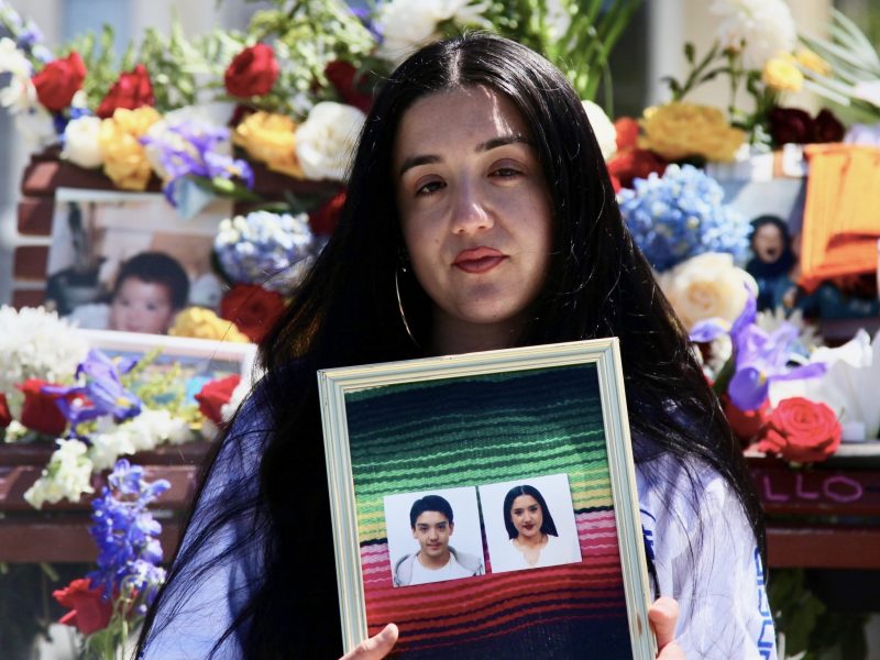 Natalia Arguello-Inglis is holding a framed picture of her and her brother, Luis Manuel Arguello-Inglis, in front of a memorial adorned with flowers and photographs.