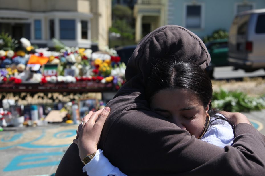 Natalia Arguello-Inglis and a friend of Luis Manuel Arguello-Inglis embrace in front of a memorial with many flowers and candles on a city street. The scene appears somber.