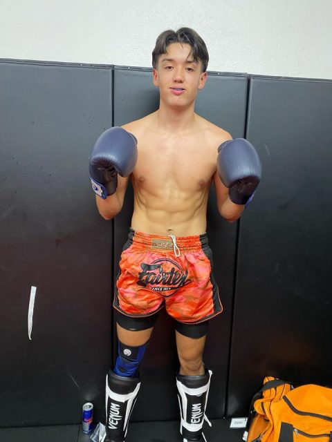 Luis Manuel Arguello Inglis with boxing gloves and shin guards stands in a gym wearing orange shorts and protective gear. An energy drink can and an orange duffel bag are on the floor beside him.