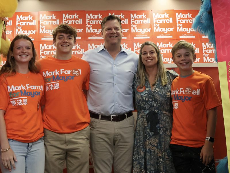 A group of five people stand together smiling, with four of them wearing orange "Mark Farrell for Mayor" shirts. They are in front of a wall covered in similar campaign signs.