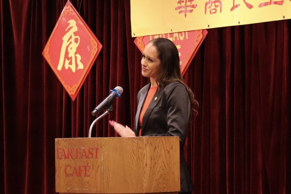 A woman in a dark suit speaks at a podium with "Far East Cafe" on it, in front of red velvet curtains and Chinese decorative ornaments.