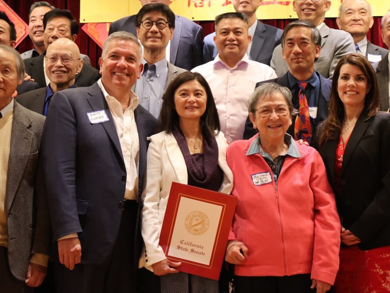 A group of people posing for a photo, with one woman in the center holding a framed "California State Senate" certificate. They are dressed in formal and semi-formal attire.