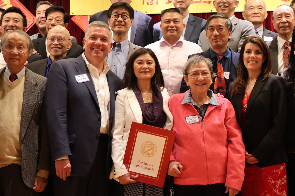 A group of people posing for a photo, with one woman in the center holding a framed "California State Senate" certificate. They are dressed in formal and semi-formal attire.