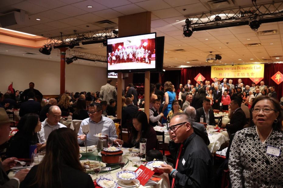 一大群人在宴会厅里或坐或站，舞台和背景屏幕上显示着一张合影。桌子上摆放着一盘盘食物。