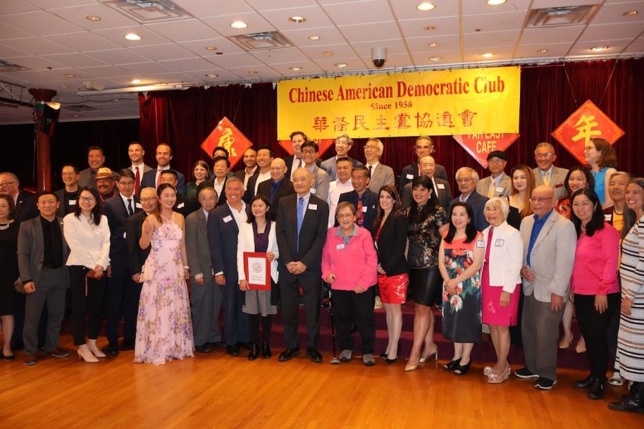 A group photo of individuals standing on a stage under a banner that reads "Chinese American Democratic Club Since 1958" in English and Chinese. The setting appears to be an indoor event or gathering.