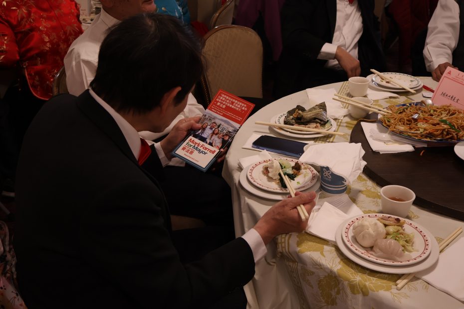 餐桌旁，一个身着正装的人拿着一本小册子，一边用筷子夹菜。餐桌上摆放着各种碗碟。