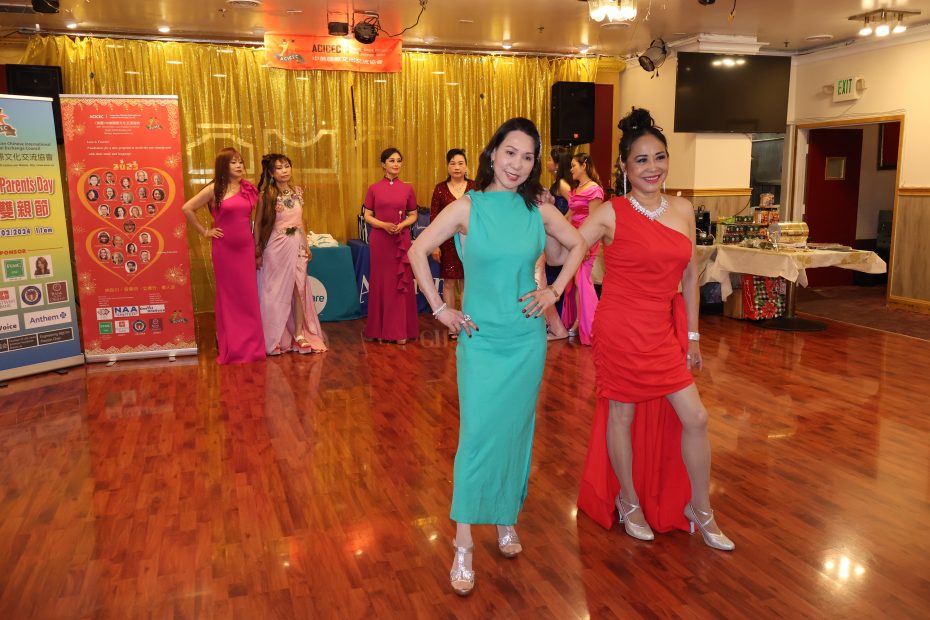 两位身着优雅礼服的女士在舞池中翩翩起舞，周围还有其他身着类似服饰的女士。背景是一个光线充足的活动大厅内的横幅、餐桌和金色窗帘。