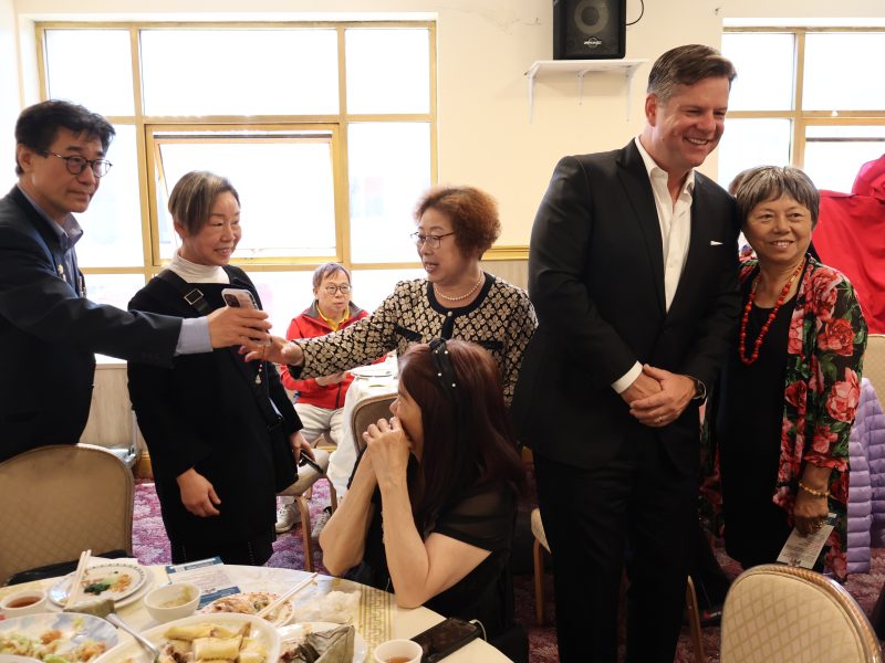 A group of people smiling and greeting each other in a room with a table set with food in the foreground and windows in the background.