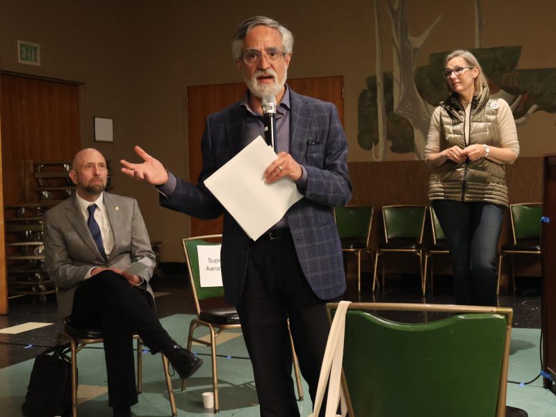 A man holding a microphone and papers speaks to an audience, with two individuals, one seated and one standing, in the background.