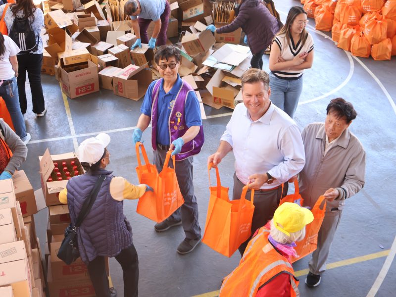 People are distributing orange bags filled with items at a community event. Boxes and other bags are visible in the background.