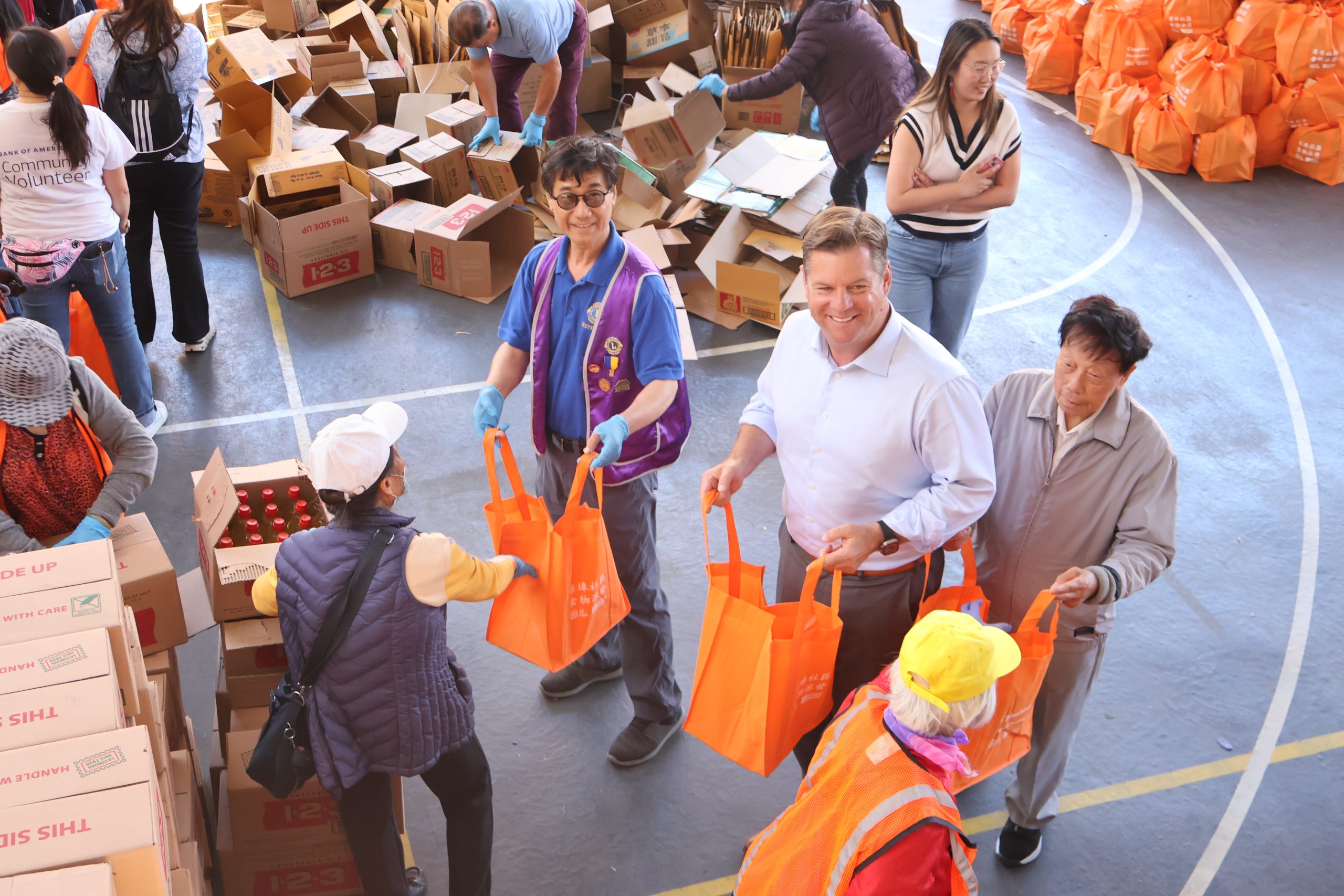 People are distributing orange bags filled with items at a community event. Boxes and other bags are visible in the background.