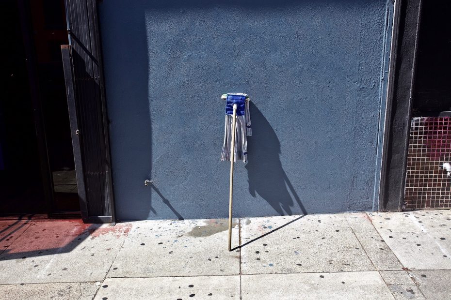 A cleaning mop with a long handle rests against a blue-painted wall on a sidewalk. The mop casts a shadow on the wall and pavement.