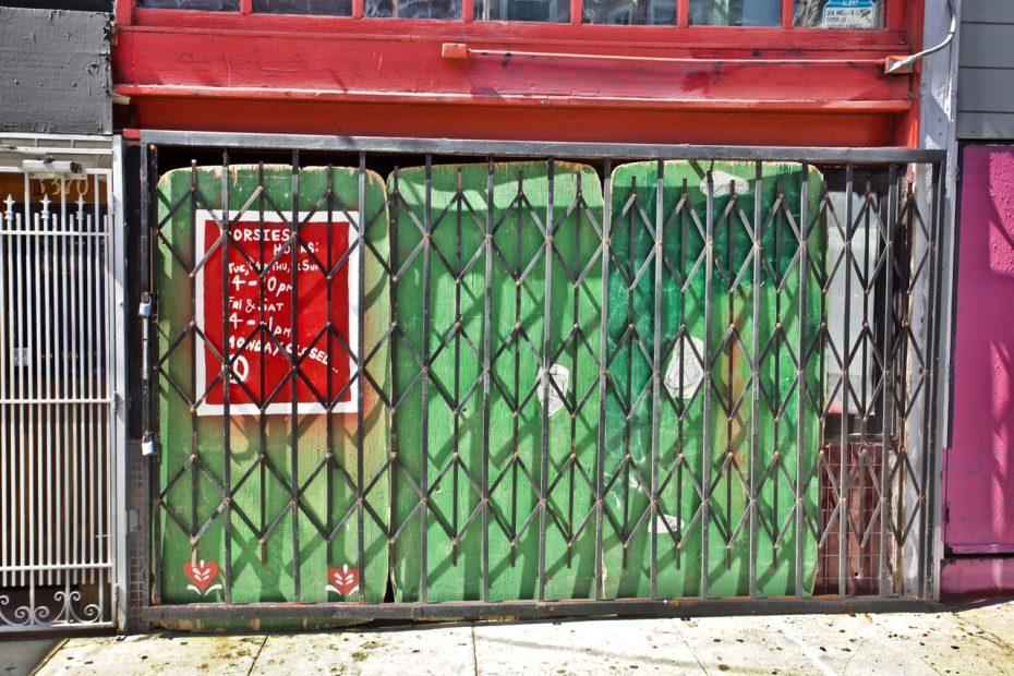 A storefront with a metal security gate and a green painted door. A red sign with white text is on the left side of the door.