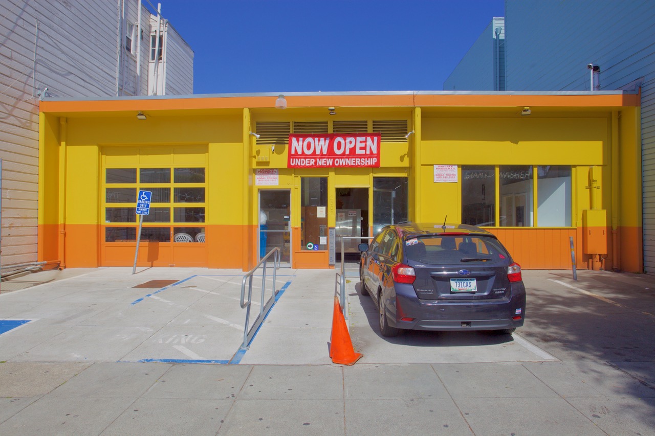 A small storefront with a bright yellow exterior and an orange trim has a "Now Open Under New Ownership" sign. A gray car is parked in front, and there is a single orange cone by the entrance.