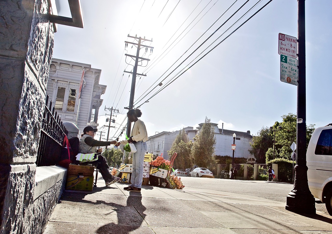 A street vendor sitting on a corner sells produce to a passerby on a sunny day, with a residential building and a "No Parking" sign visible in the background.