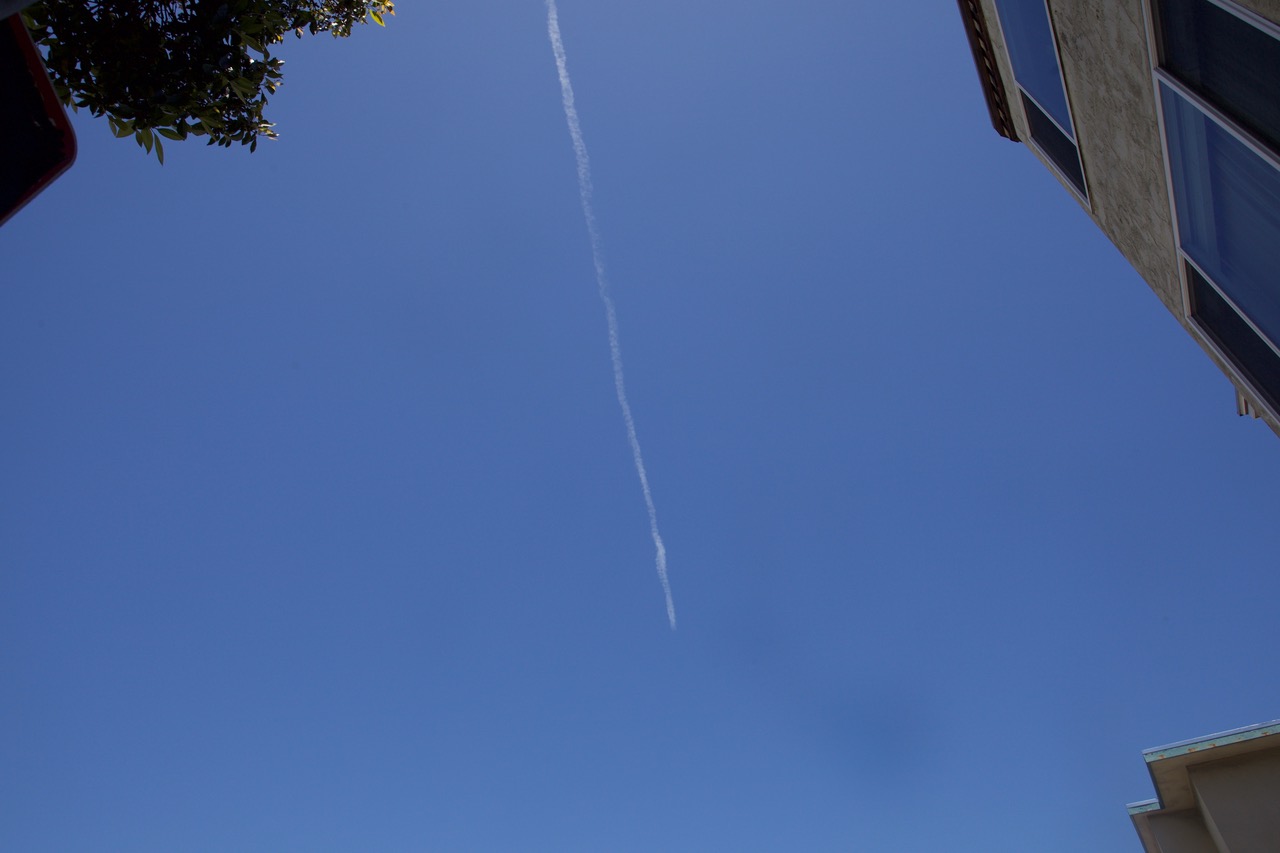 A single contrail stretches diagonally across a clear blue sky, with parts of two buildings and a tree visible at the edges.