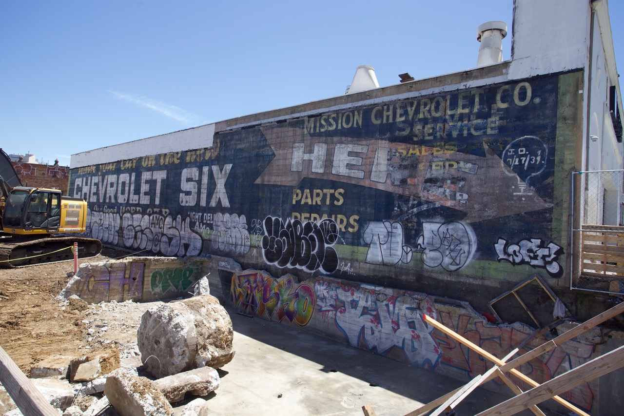 An old, faded Chevrolet advertisement billboard on a building wall with graffiti. Construction equipment and debris are present in the foreground.