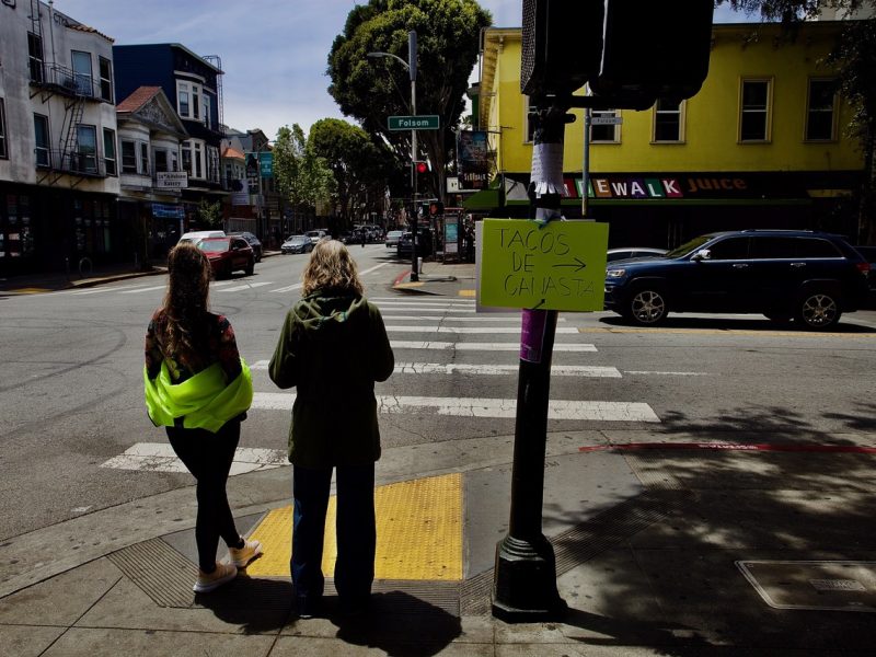 Two people stand at a street corner next to a pole with a yellow sign that reads "Tacos de Guiso" with an arrow pointing right. Buildings and parked cars are visible in the background.