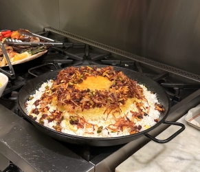 A round dish of baked rice topped with caramelized onions and pistachios sits on a stovetop, with various utensils and food items on the adjacent counter.